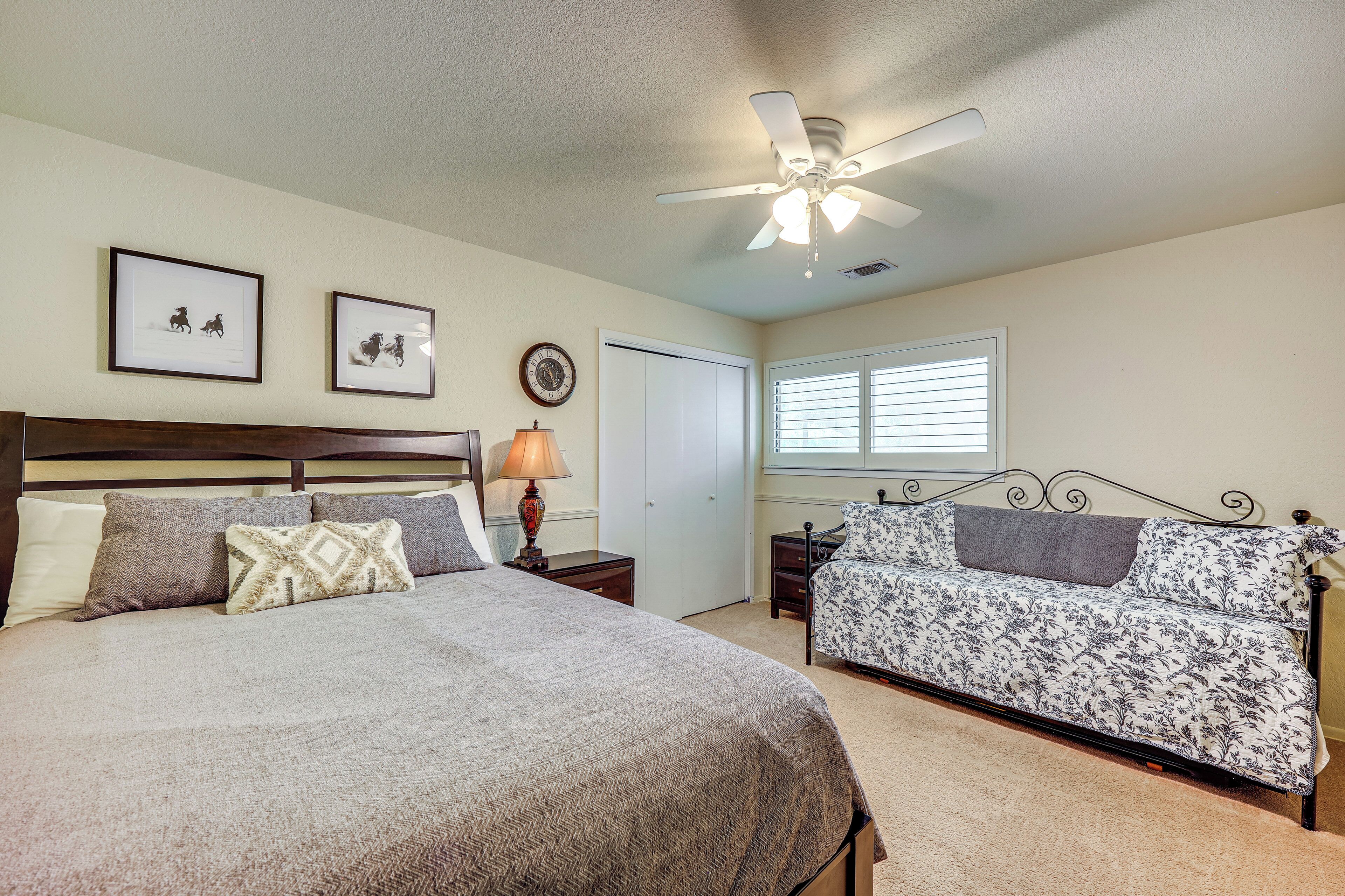A bedroom with a large bed, a smaller sofa, a ceiling fan, framed pictures on the wall, and a window with blinds.