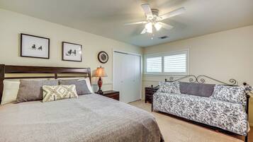 A bedroom with a large bed, a smaller sofa, a ceiling fan, framed pictures on the wall, and a window with blinds.