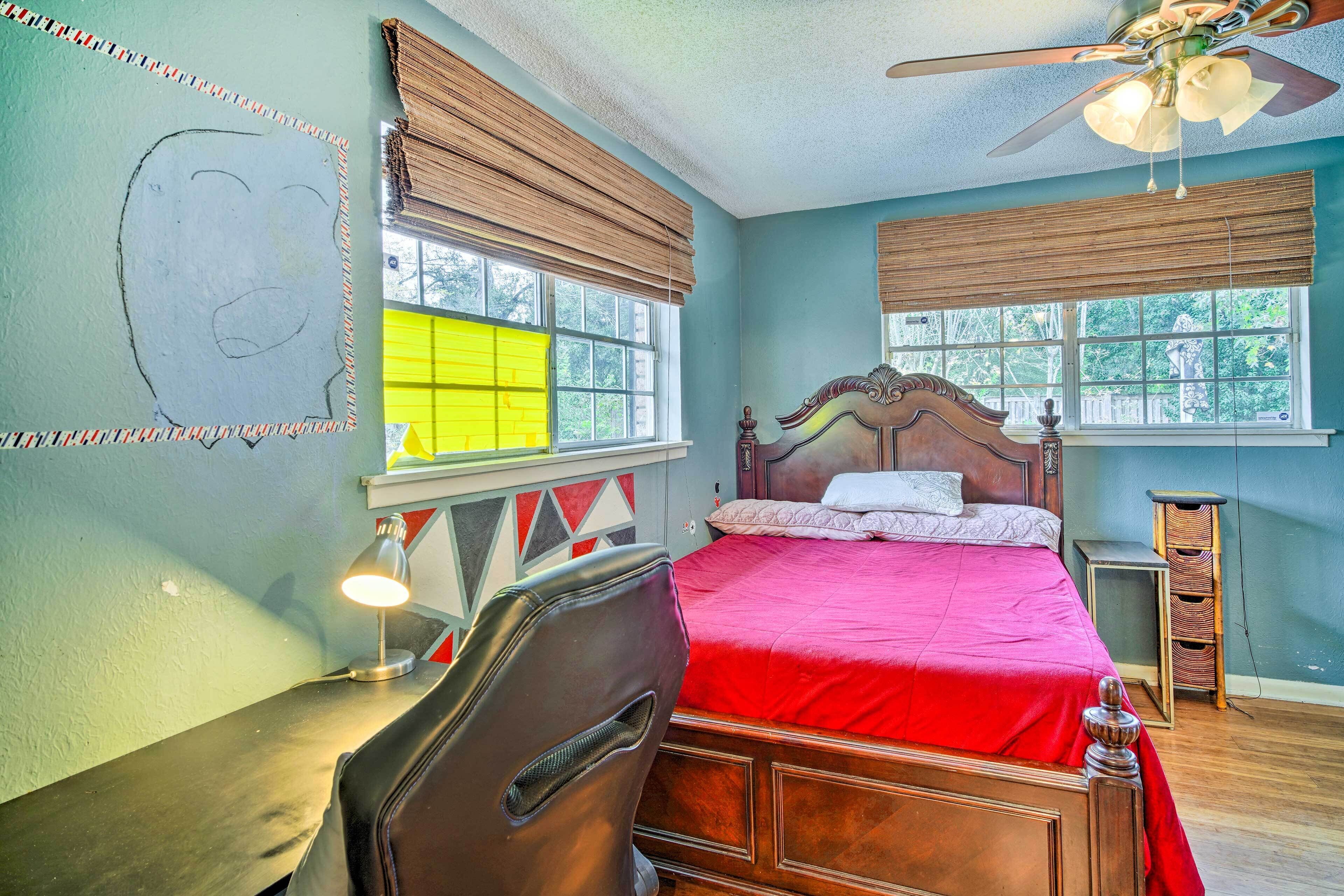 Une chambre à coucher comprenant un lit, un bureau, une chaise, un ventilateur de plafond et des fenêtres équipées de stores.