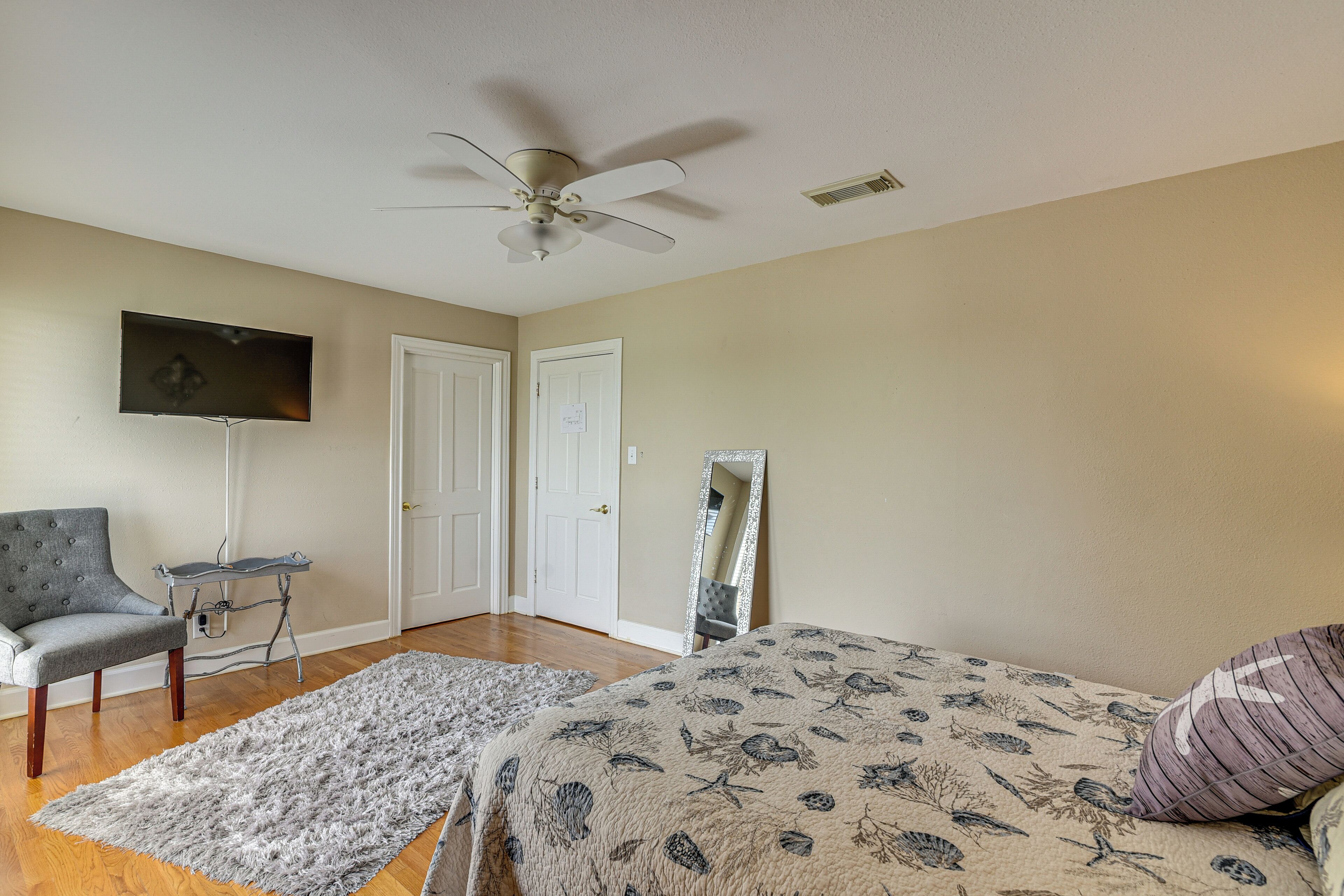 A bedroom with a bed, a chair, a TV, a ceiling fan, and a mirror.