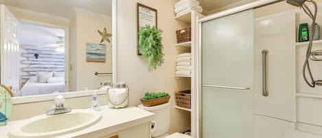 A bathroom with a sink, toilet, and shower. There are towels, a plant, and a framed picture on the wall.