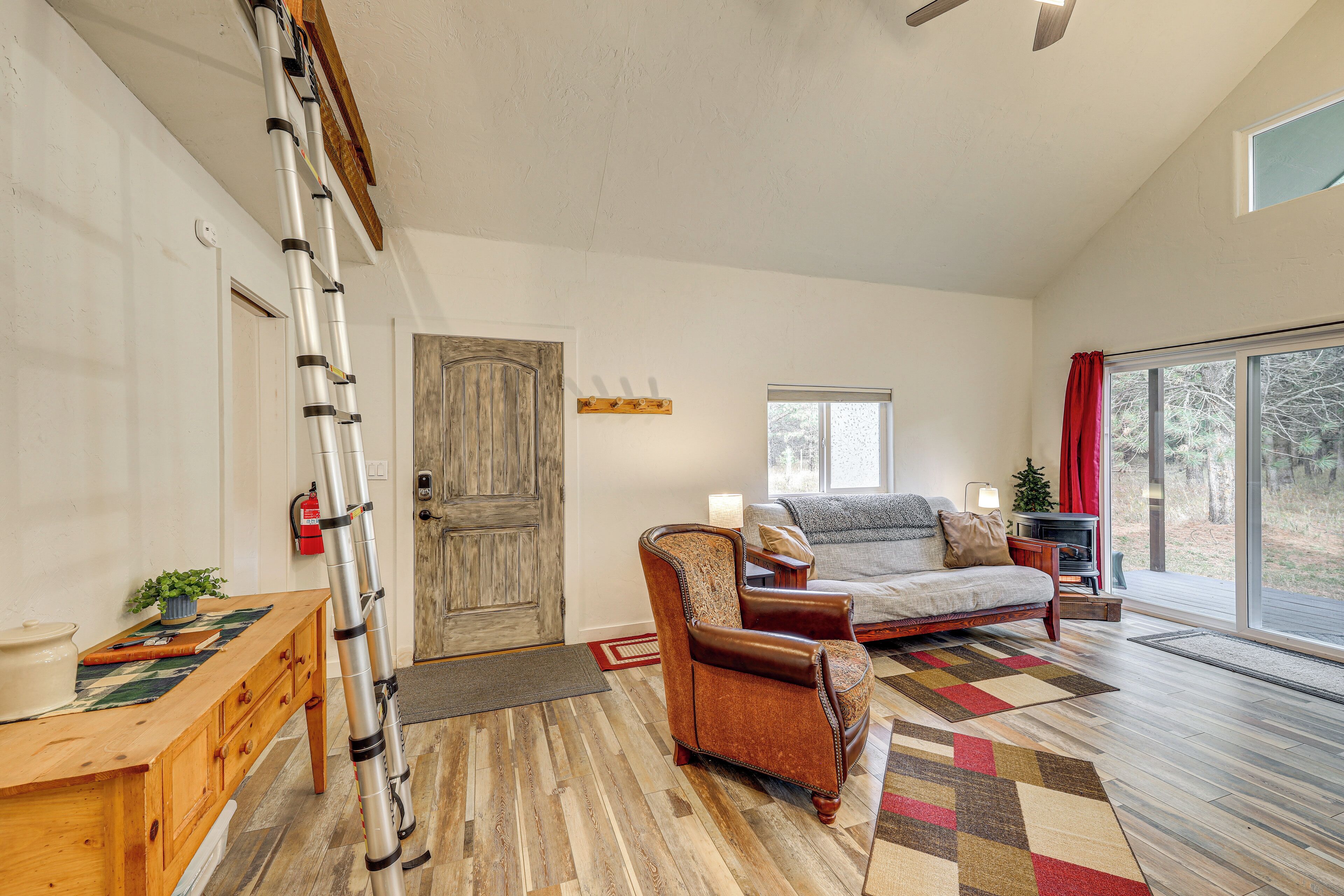 A living room with a wooden floor, a sofa, a chair, a ladder, a wooden door, and a window.