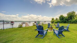 Eine Rasenfläche mit Adirondack-Stühlen und einer Feuerstelle, mit Blick auf einen See mit Booten und Stegen.