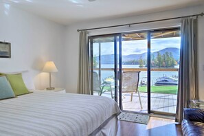 A hotel room with a bed, a nightstand, a lamp, and a view of a lake and mountains through a sliding glass door. - Bolton Landing Suite w/ Deck, Lake George Access (Bolton Landing)