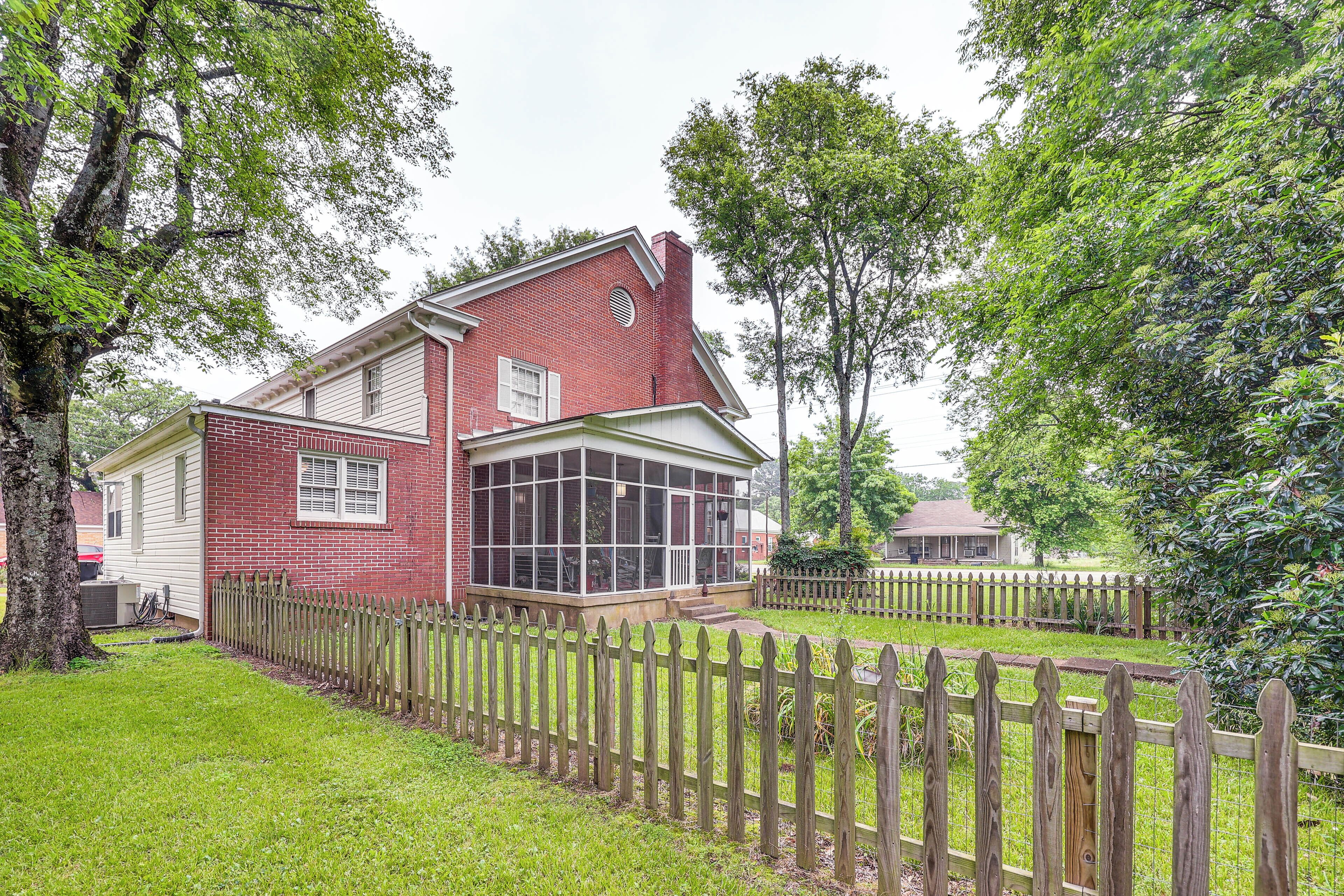 A two-story house with a large glass windowed porch, surrounded by a wooden fence and lush greenery.