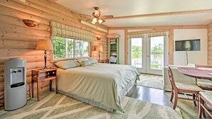 A log cabin bedroom with a bed, bedside tables, a heater, a fan, a TV, and a dining area with chairs. - Rock River Access! Peaceful Horicon Studio (Horicon)