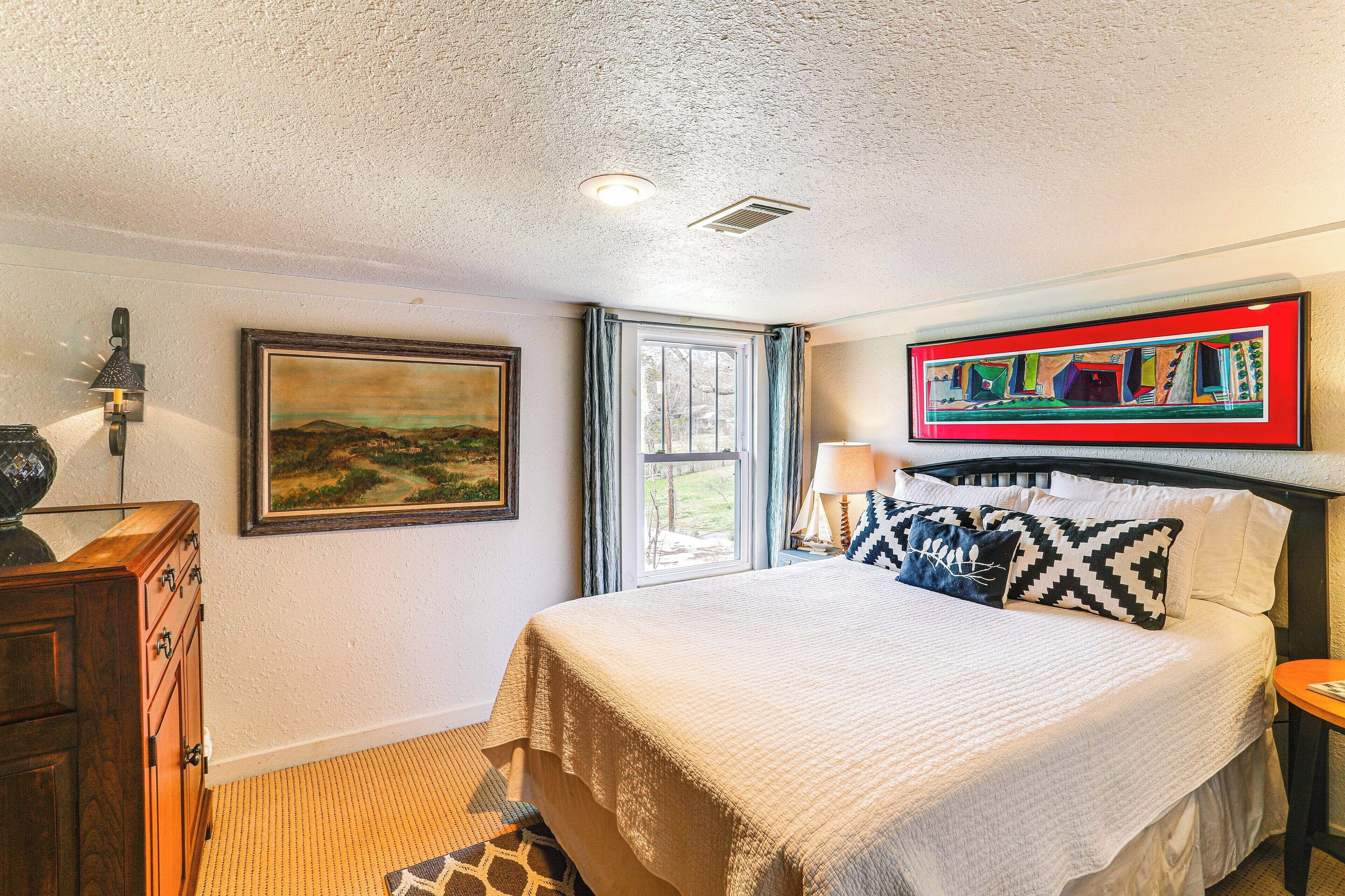 A bedroom with a bed, a nightstand, a lamp, a painting, a window with curtains, and a wall-mounted picture frame.