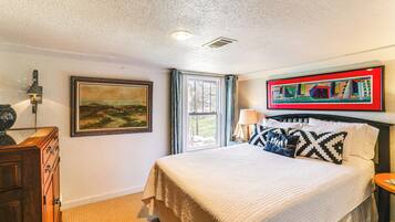 A bedroom with a bed, a nightstand, a lamp, a painting, a window with curtains, and a wall-mounted picture frame.