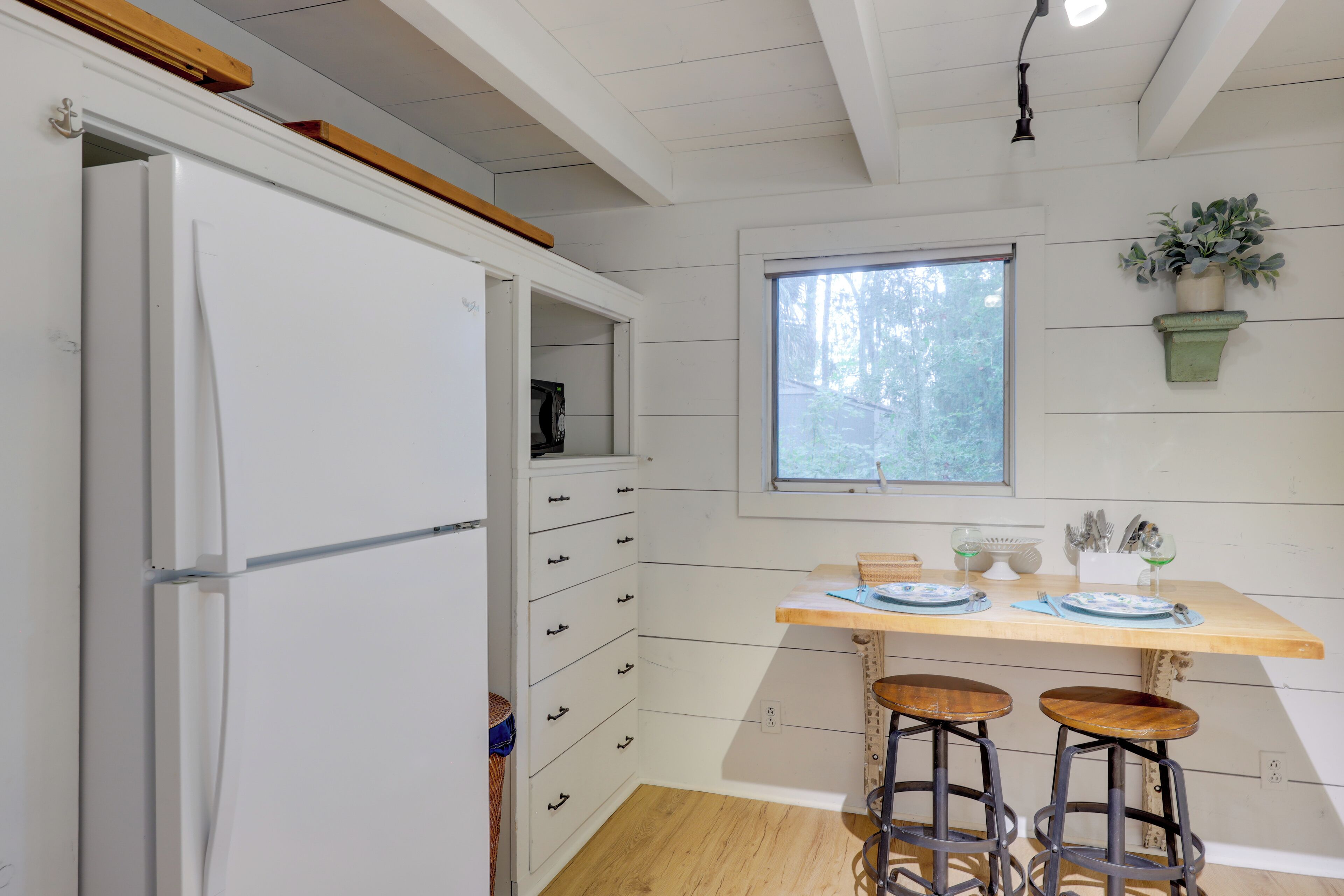 Una cocina con un refrigerador blanco, encimera de madera, dos taburetes y una ventana con vista a árboles.