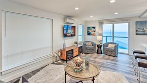 A spacious living room with a large window, a television, a wooden cabinet, a patterned sofa, and a glass coffee table. - Tacoma 'dash Point Beach Cabin' w/ Balcony! (Tacoma)