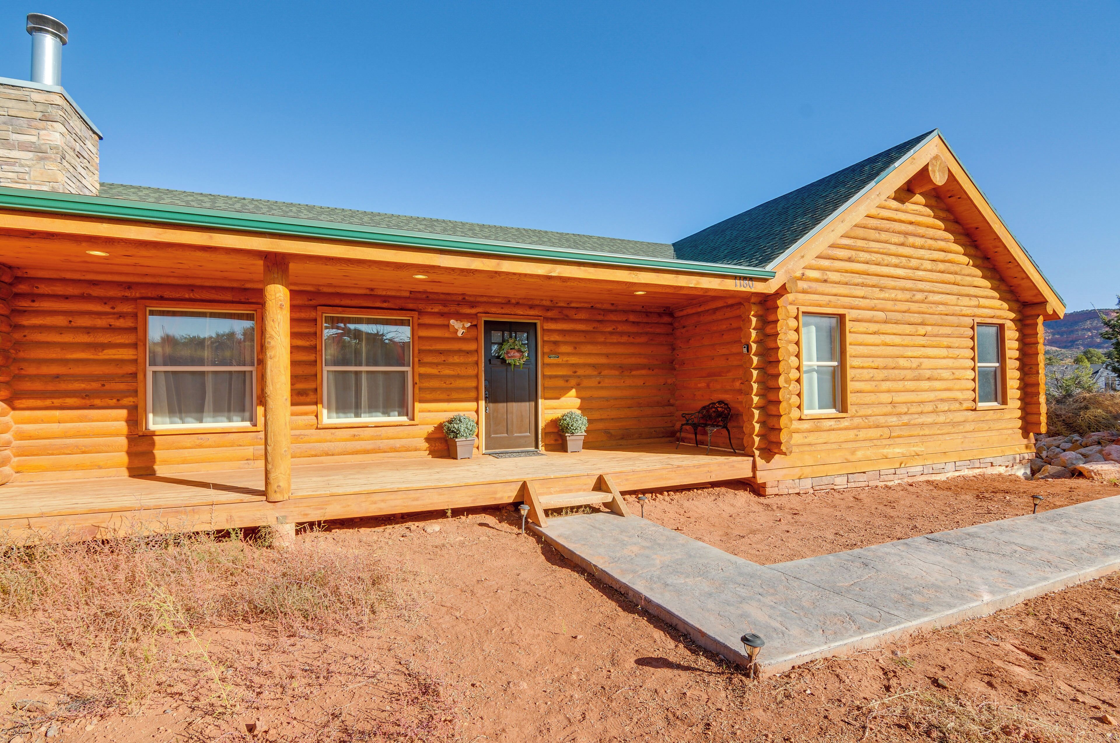 A log cabin with a green roof, a chimney, and a porch with potted plants.