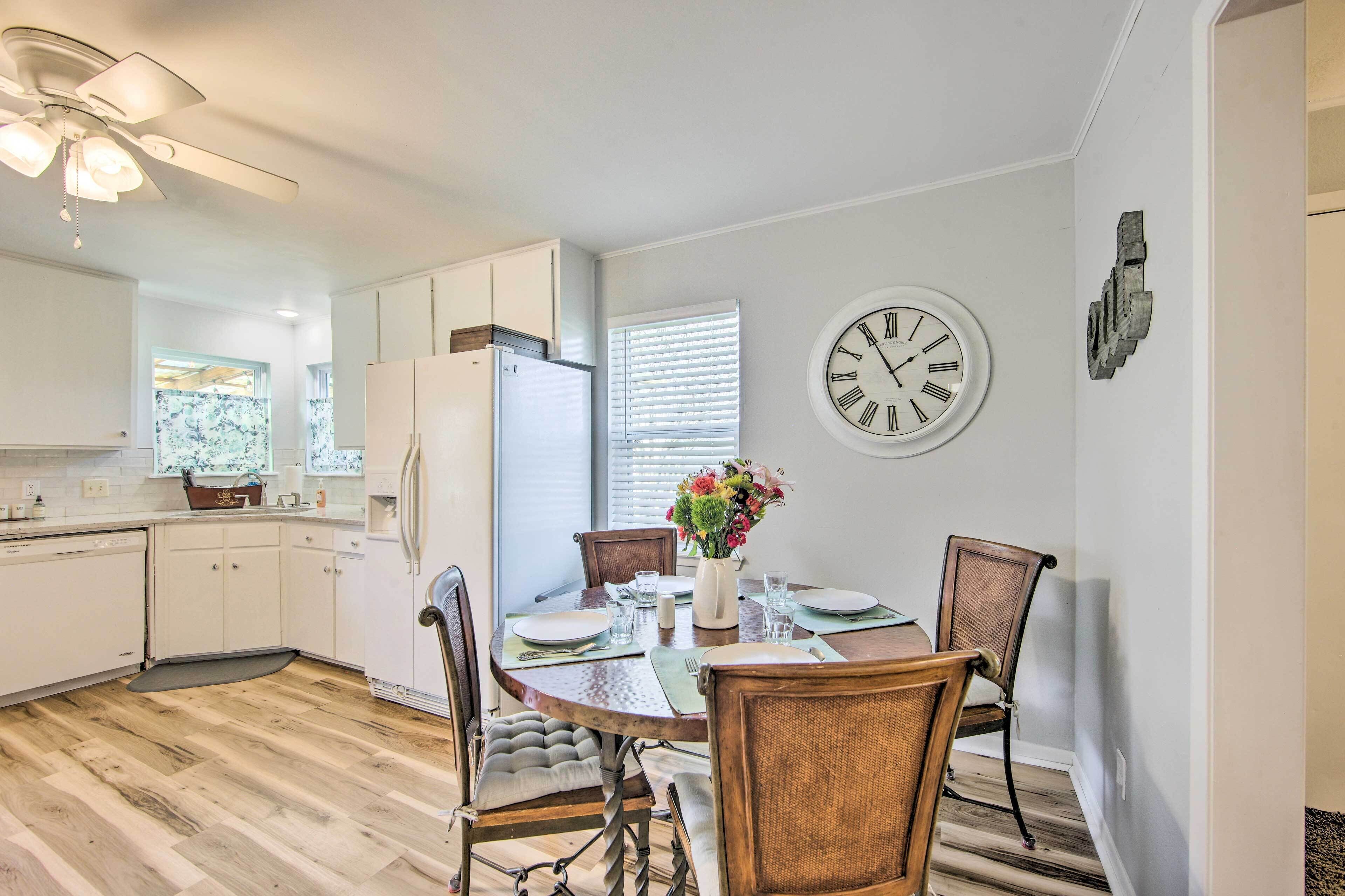 A modern kitchen with white cabinets, a dining table with chairs, a clock on the wall, and a window with blinds.