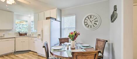 A modern kitchen with white cabinets, a dining table with chairs, a clock on the wall, and a window with blinds.