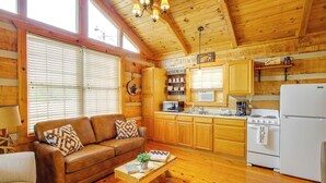 A cozy cabin interior with a wooden floor, a brown leather sofa, a wooden coffee table, a kitchen area with wooden cabinets, a stove, and a refrigerator. - Rivers View - Cherokee Lake Cabin w/ Fire Pit! (Bean Station)