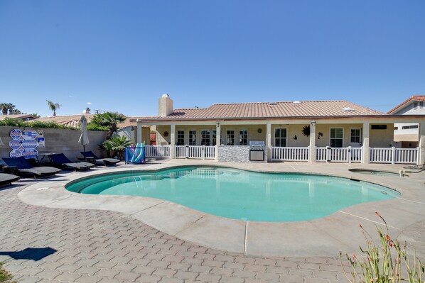 A swimming pool with a curved design, a covered patio area with lounge chairs, and a white fence surrounding the property.