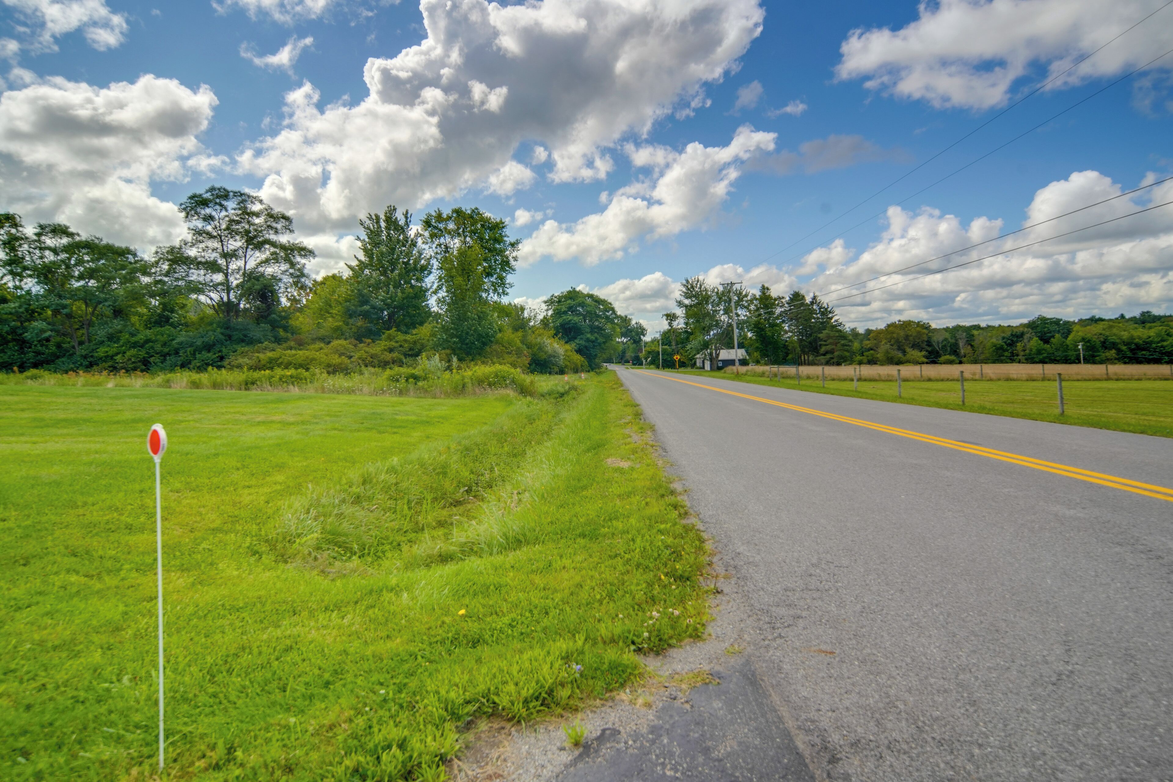 A rural road with a grassy field on one side and a fence on the other.