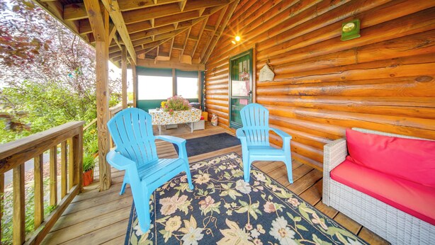 Una baita di legno con una terrazza in legno, due poltrone Adirondack blu, un divano in vimini con cuscini rossi e un tappeto floreale.