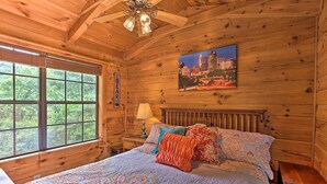A wooden cabin bedroom with a bed, a nightstand, a lamp, and a cityscape painting on the wall. - Quaint Log Cabin w/ Views Near West Jefferson (Warrensville)
