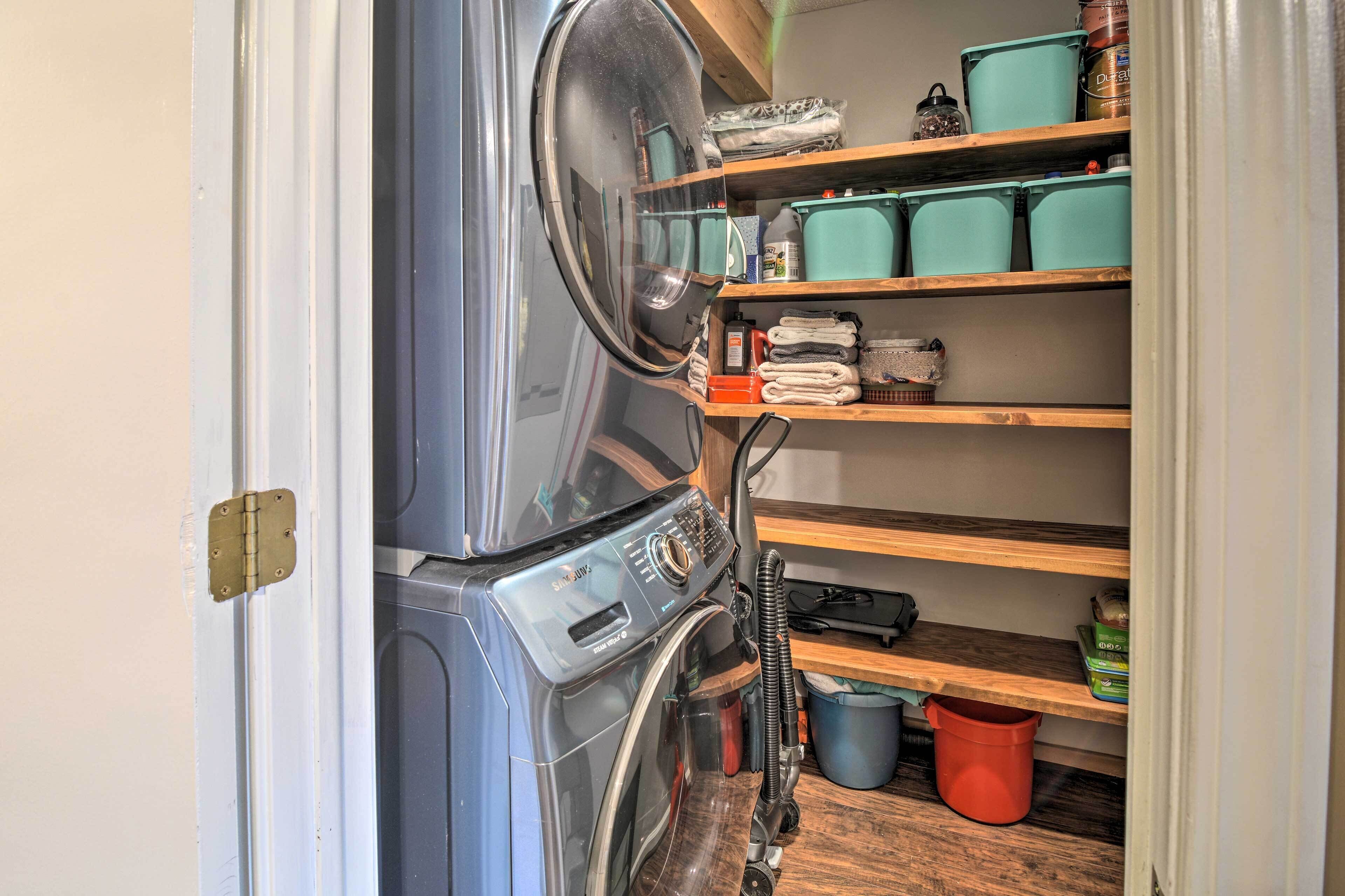 A compact laundry area with a washing machine, shelves with storage bins, and a red bucket.