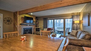 A cozy living room with a wooden dining table, a fireplace, a sofa, and a large window with a view of trees. - Cozy Bellaire Condo w/ Balcony - 3 Mi to Skiing! (Bellaire)