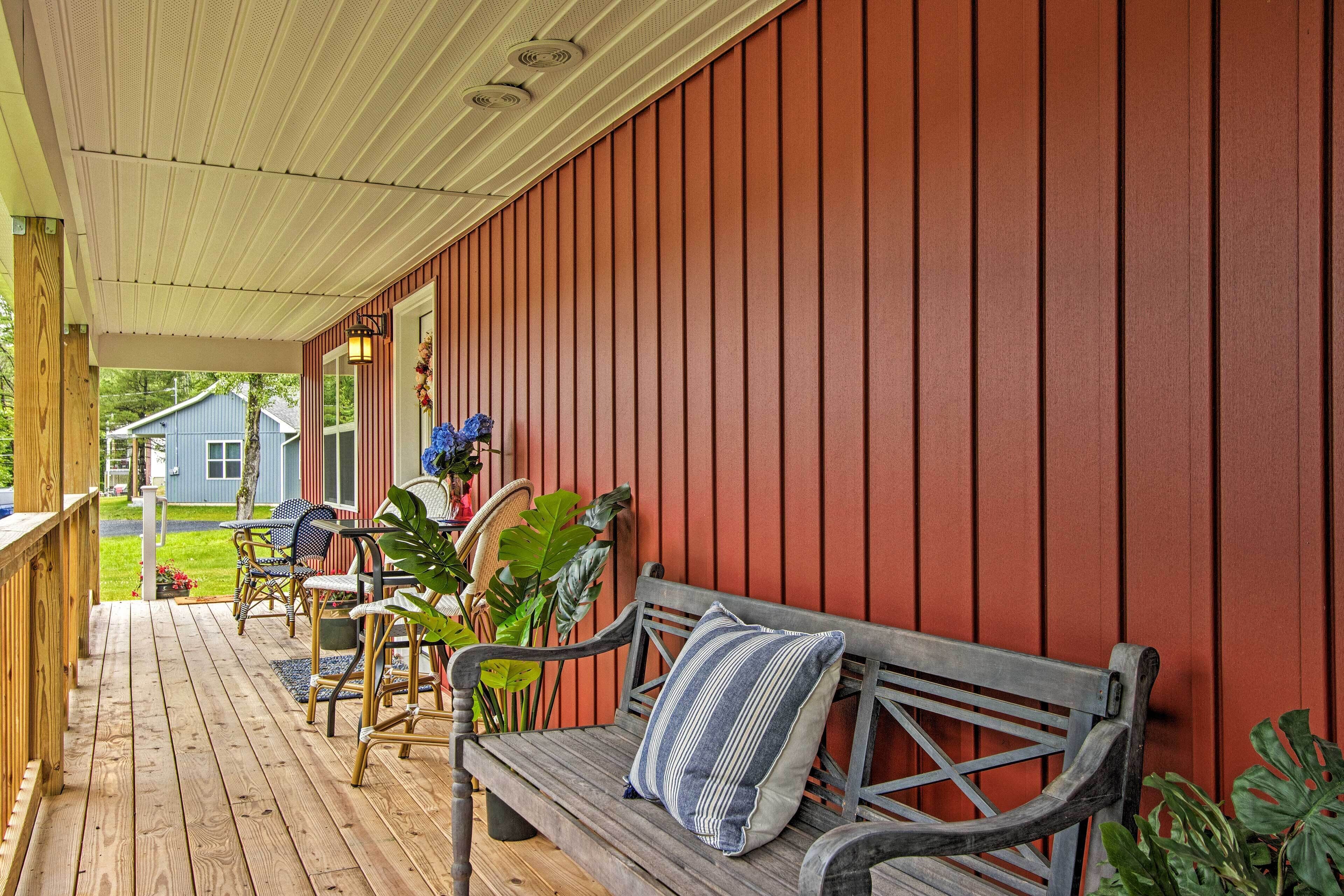 A wooden deck with a bench and chairs, a red wall, and a house in the background.