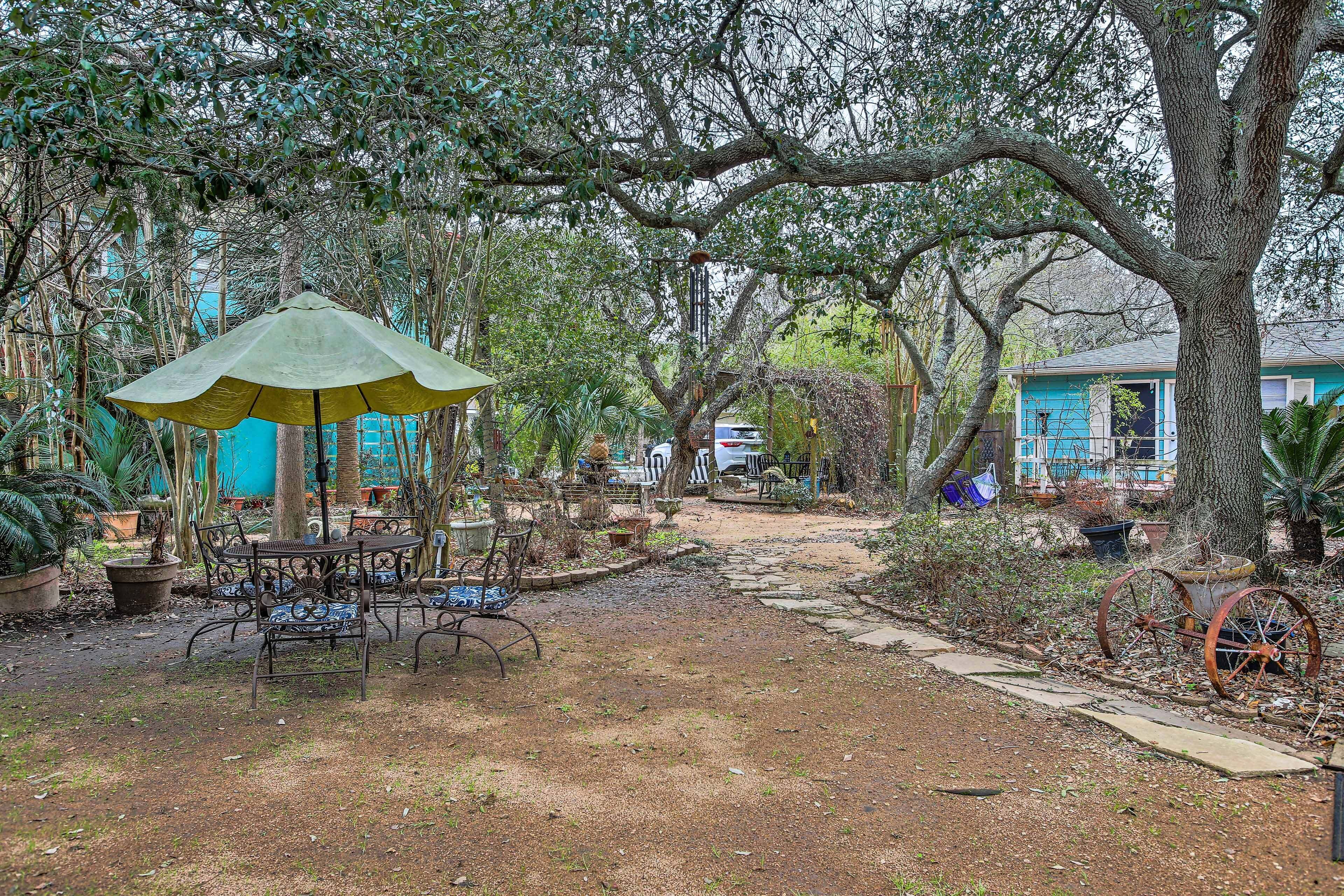 A rustic outdoor area with a table and chairs under a green umbrella, surrounded by trees and a pathway leading to a house.