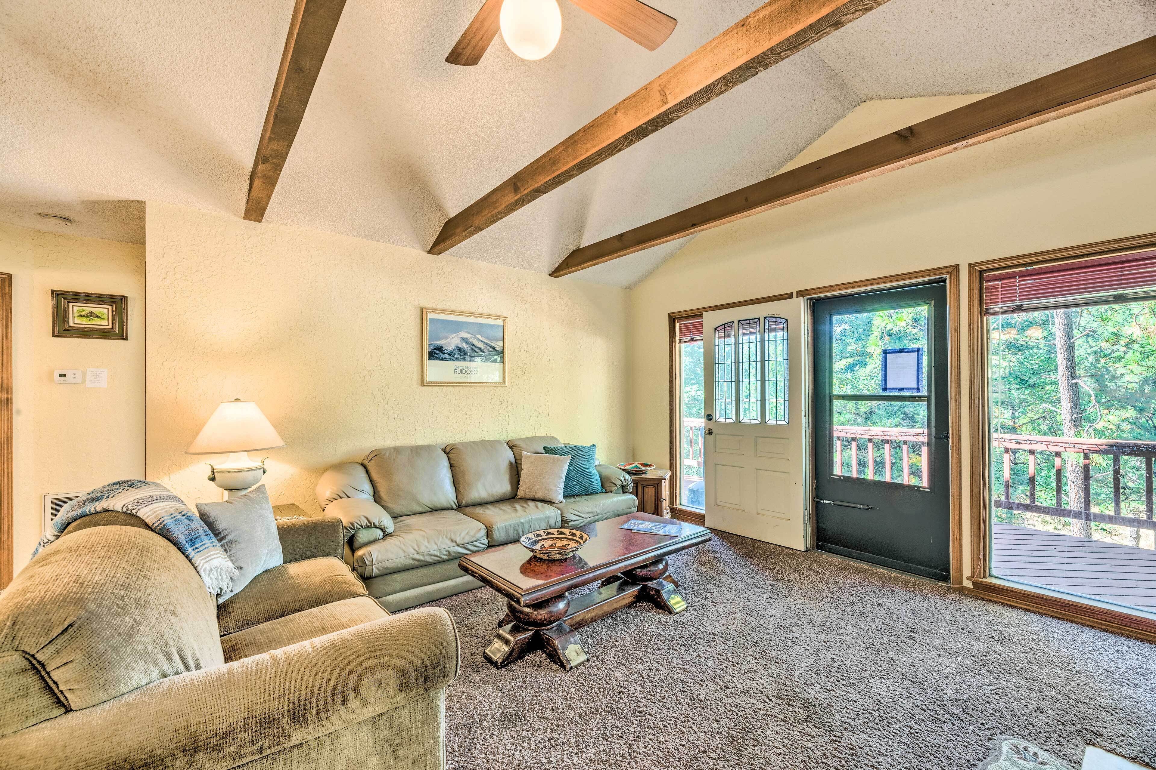 A cozy living room with a sofa, a coffee table, and a lamp. There is a door leading to a balcony with a view of trees.