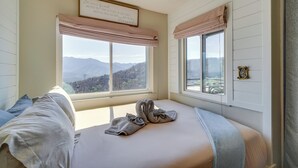 A bedroom with a bed, two windows with blinds, a view of mountains, and a framed sign on the wall.
