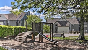 A playground with a slide and climbing structure, surrounded by a grassy area and a row of houses in the background.
