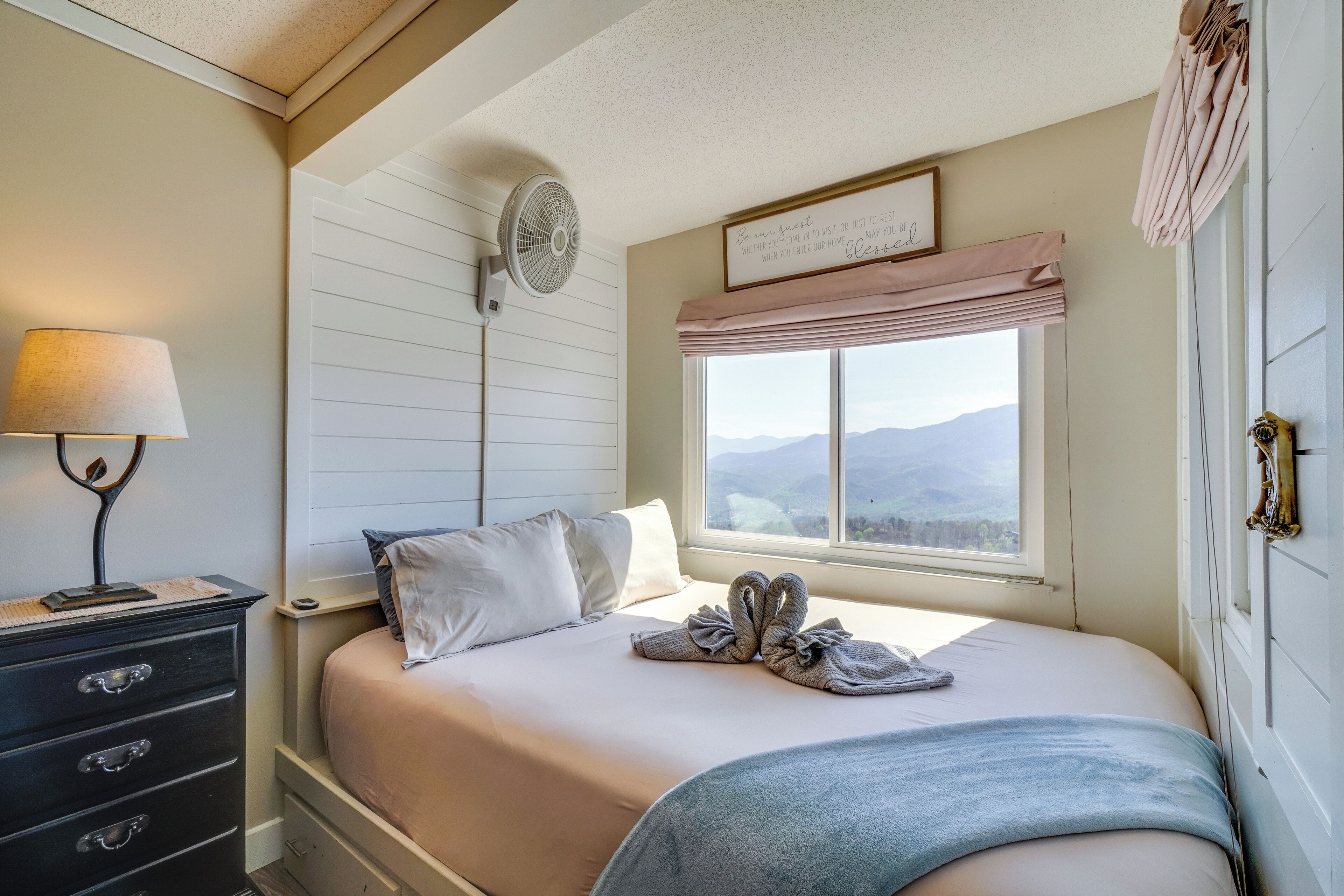 Cama bem arrumada com vista para montanhas pela janela, abajur e ventilador fixado na parede.