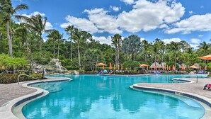 A large, curved swimming pool with a clear blue water surface, surrounded by palm trees and lounge chairs with orange umbrellas. - Alluring Naples Retreat w/ Resort Amenities! (Naples)