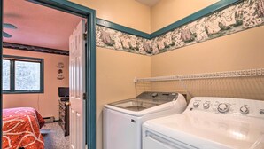 Une buanderie équipée d’un lave-linge et d’un sèche-linge, un lit recouvert d’une couvre-lit à motifs rouges et blancs, et une fenêtre donnant sur les arbres.