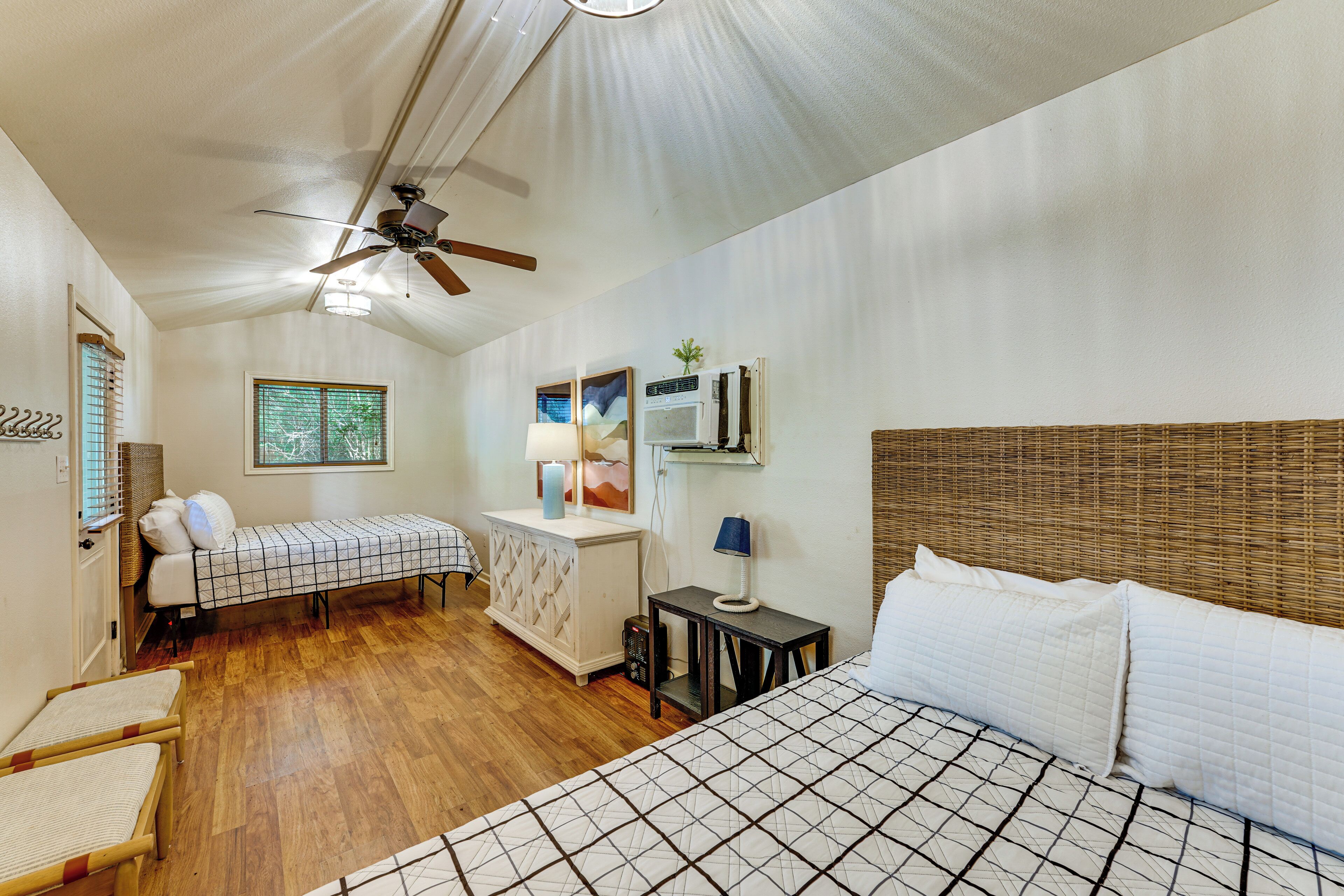 A bedroom with a large bed, a smaller bed, a desk with a lamp, a ceiling fan, and a window with blinds.
