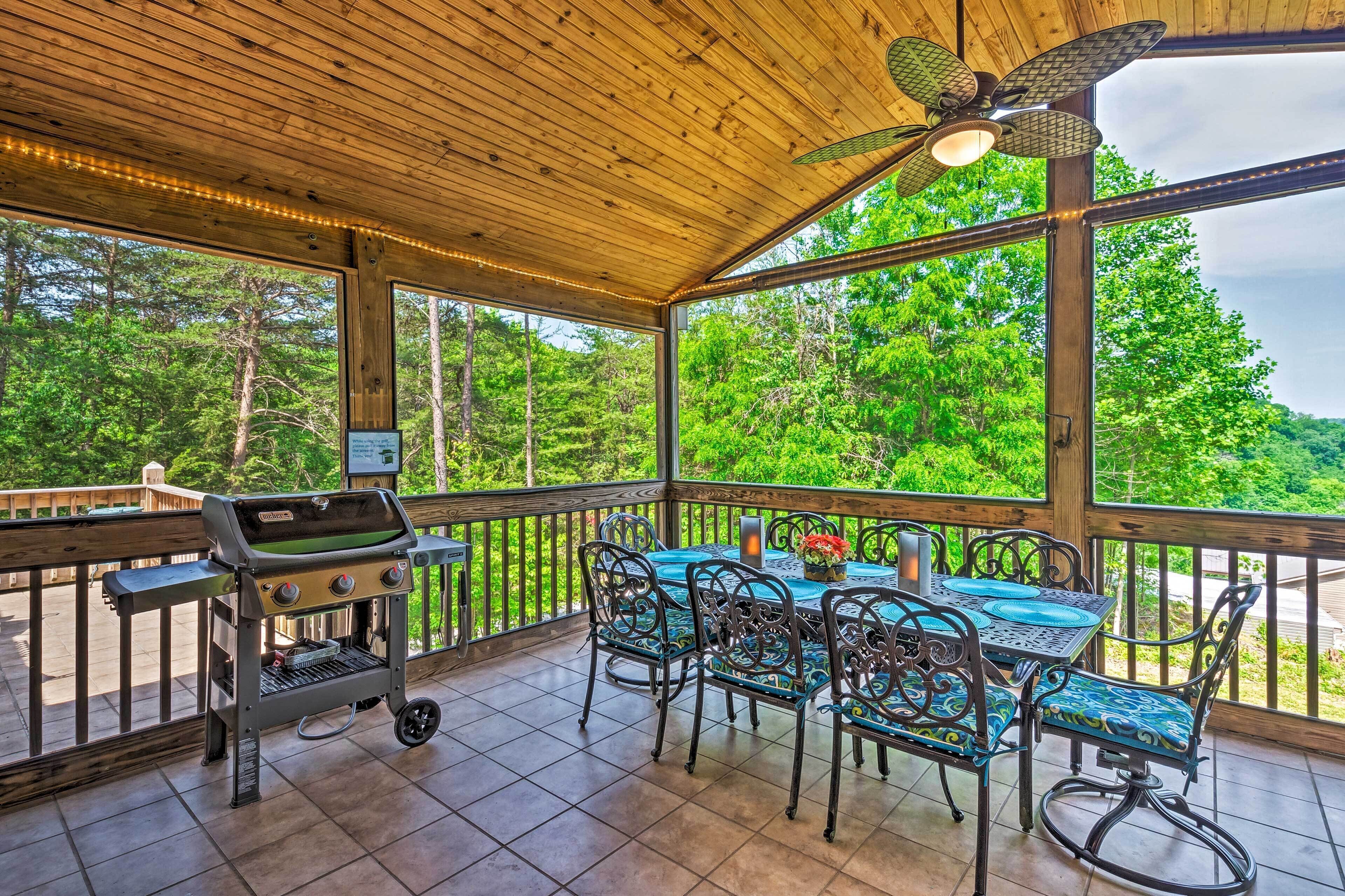 A covered outdoor area with a grill, metal chairs, and a table with a view of the forest.