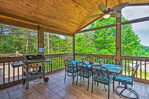 A covered outdoor area with a grill, metal chairs, and a table with a view of the forest.