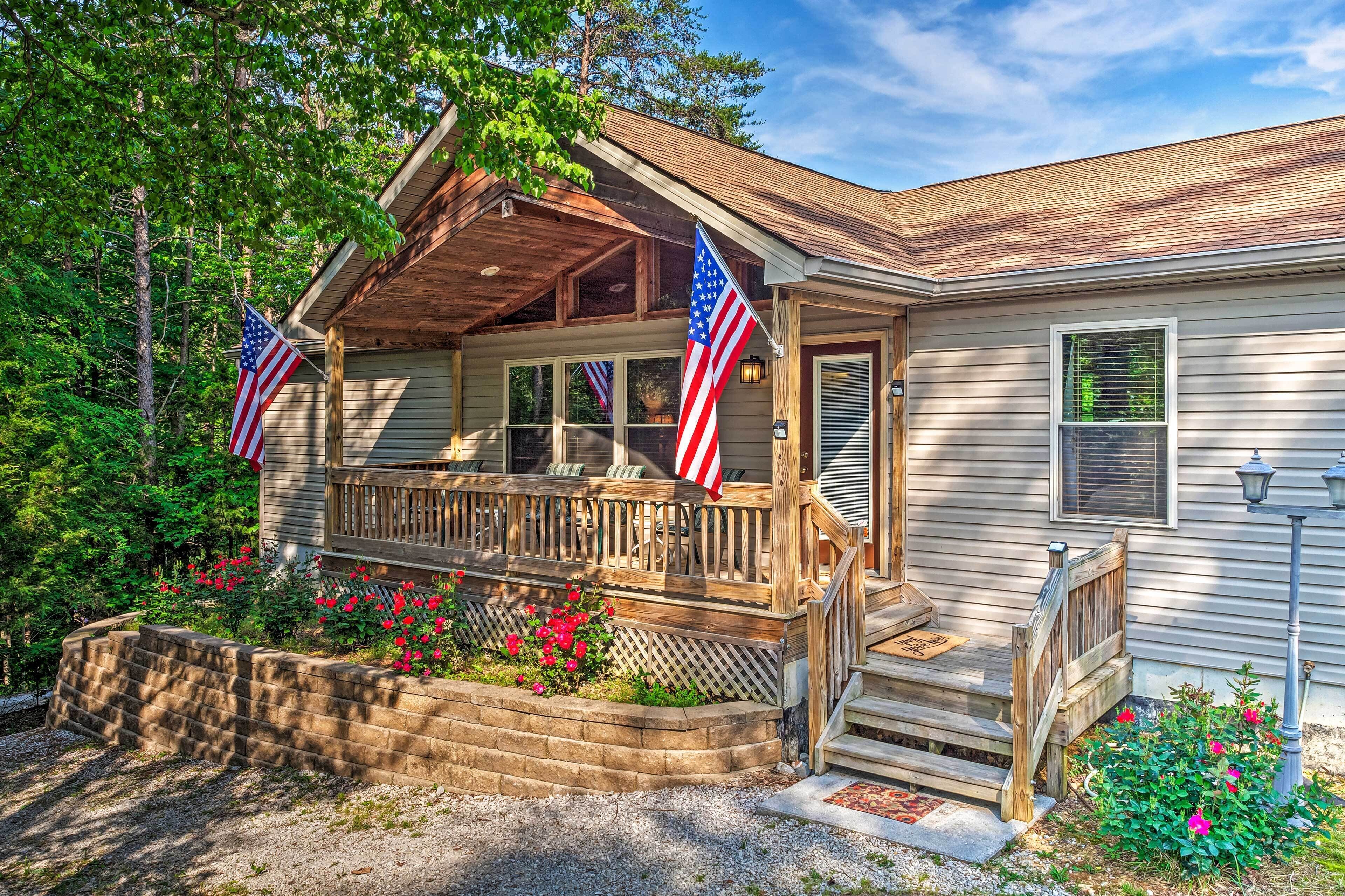 A cabin with a wooden porch, two American flags, and a landscaped garden with red flowers.