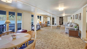 A spacious living area with a large window overlooking a deck, a cozy seating area with a sofa and chairs, and a dining table with a bowl of fruit. - Athens Vintage Fishing Cabin on Elk River! (Athens)