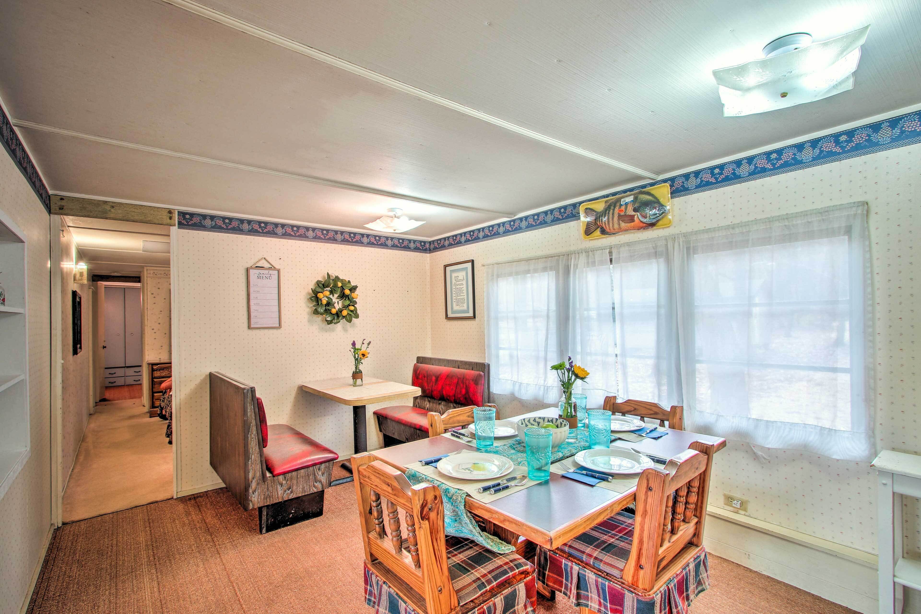 A dining area with a table set for four, surrounded by plaid-patterned chairs. There is a small table with a single chair and a vase with flowers. The room has a ceiling fan, a window with curtains, and a wall-mounted fish decoration.