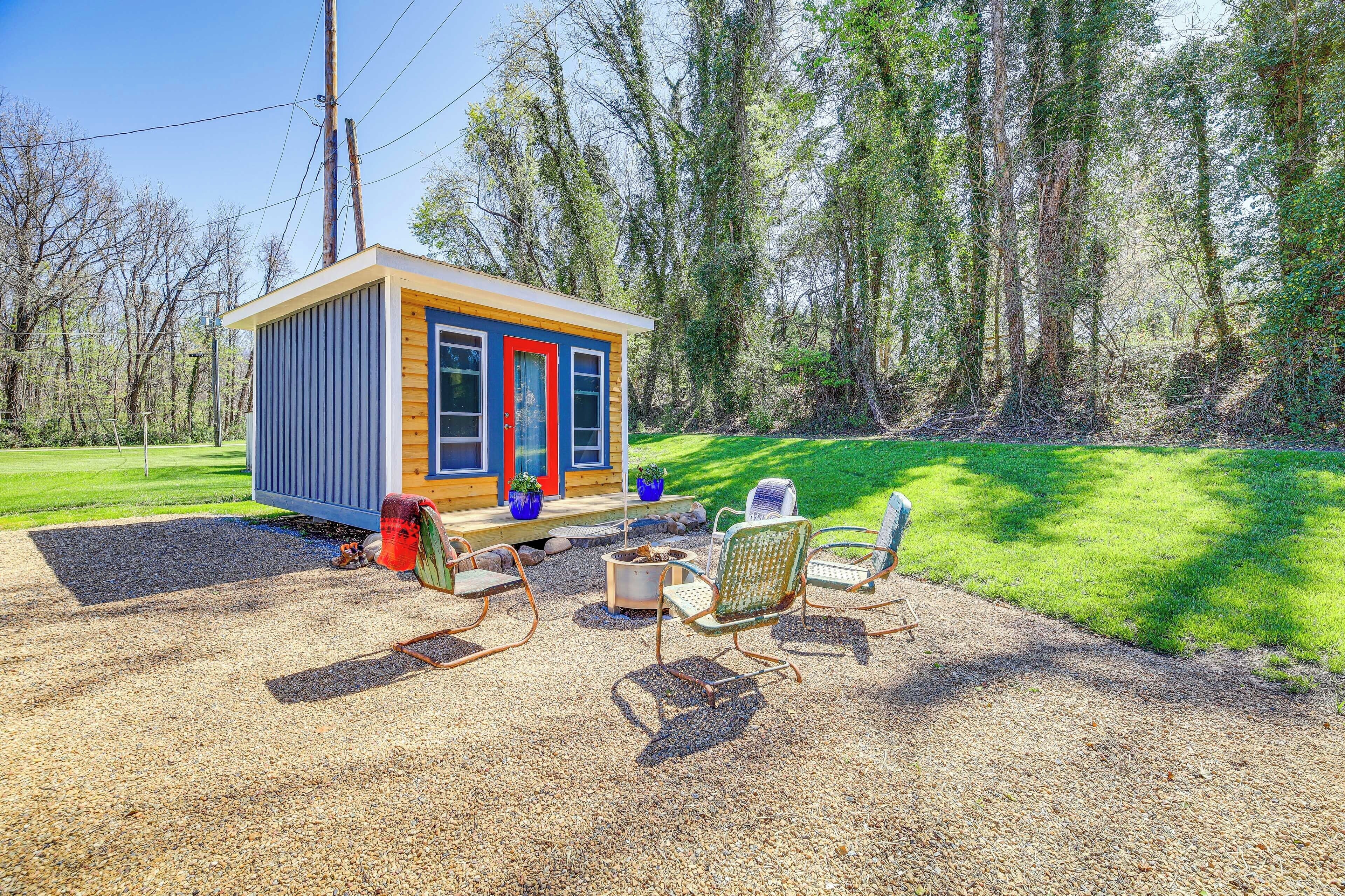 A small wooden cabin with a red door and windows, surrounded by a gravel patio with metal chairs and a fire pit.