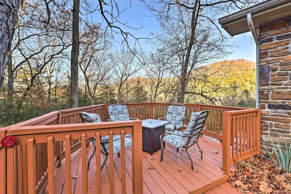 A wooden deck with outdoor furniture, a stone building, and a forested backdrop.