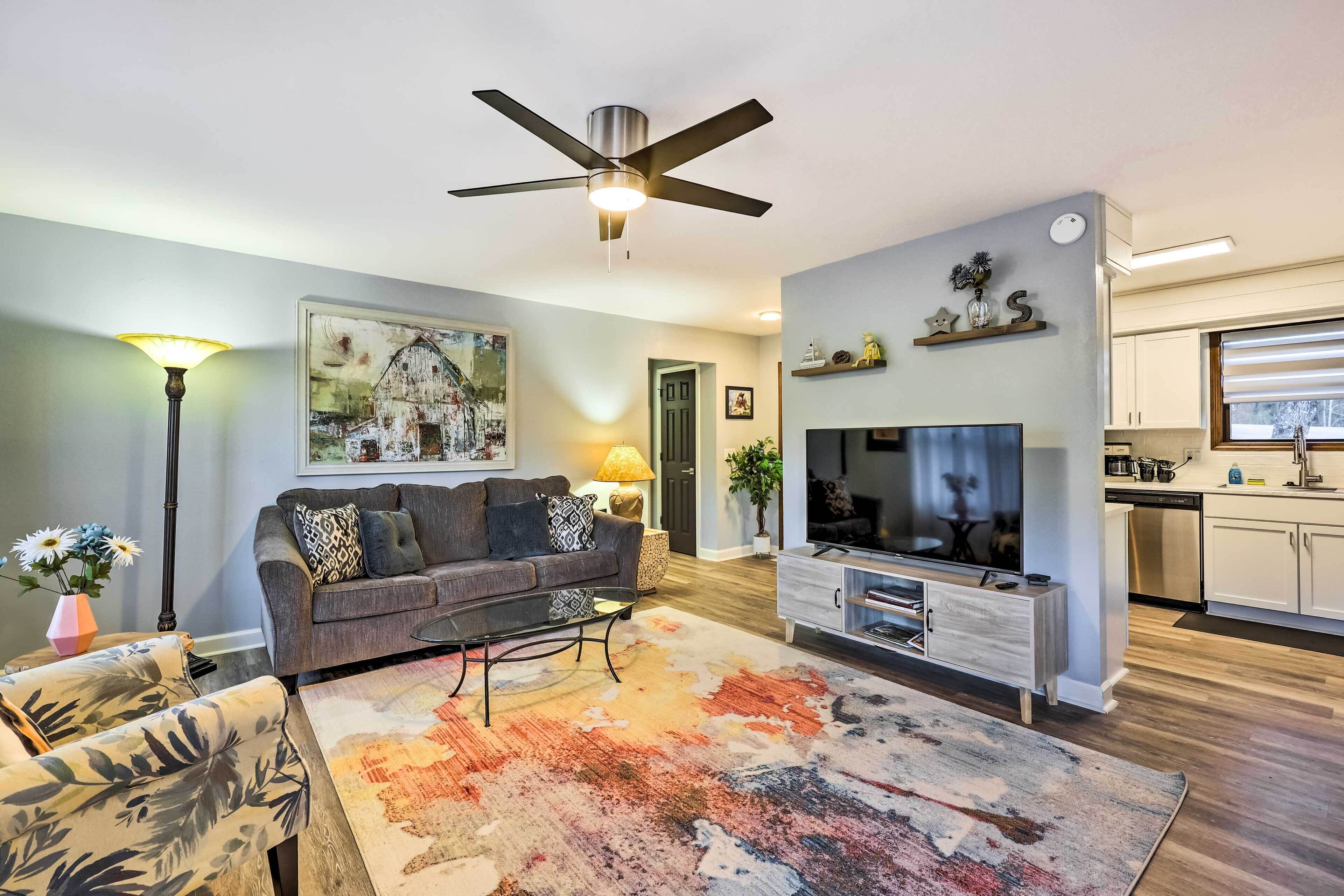 A living room with a sofa, coffee table, and TV, a ceiling fan, and a kitchen area with cabinets and appliances.