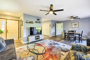 A modern living room with a flat-screen TV, a coffee table, and a sofa. There are ceiling fans, a dining area with a table and chairs, and a hallway with a door.