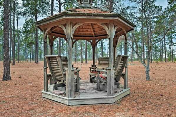 A wooden gazebo with a hexagonal roof and a central table surrounded by chairs, set in a forested area.