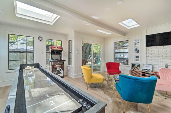A spacious room with a foosball table, a billiards table, and a billiards machine. There are two windows with blinds, a red chair, a yellow chair, and a blue chair. - Luxurious Lubbock Home: Fire Pit, Outdoor TV! (Lubbock)