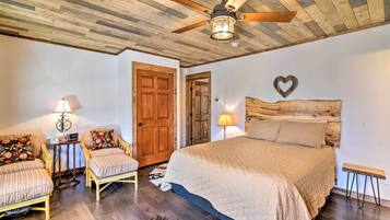 A bedroom with a wooden ceiling, a bed with a quilted cover, two upholstered chairs with floral cushions, a wooden headboard, and a wall-mounted heart decoration.