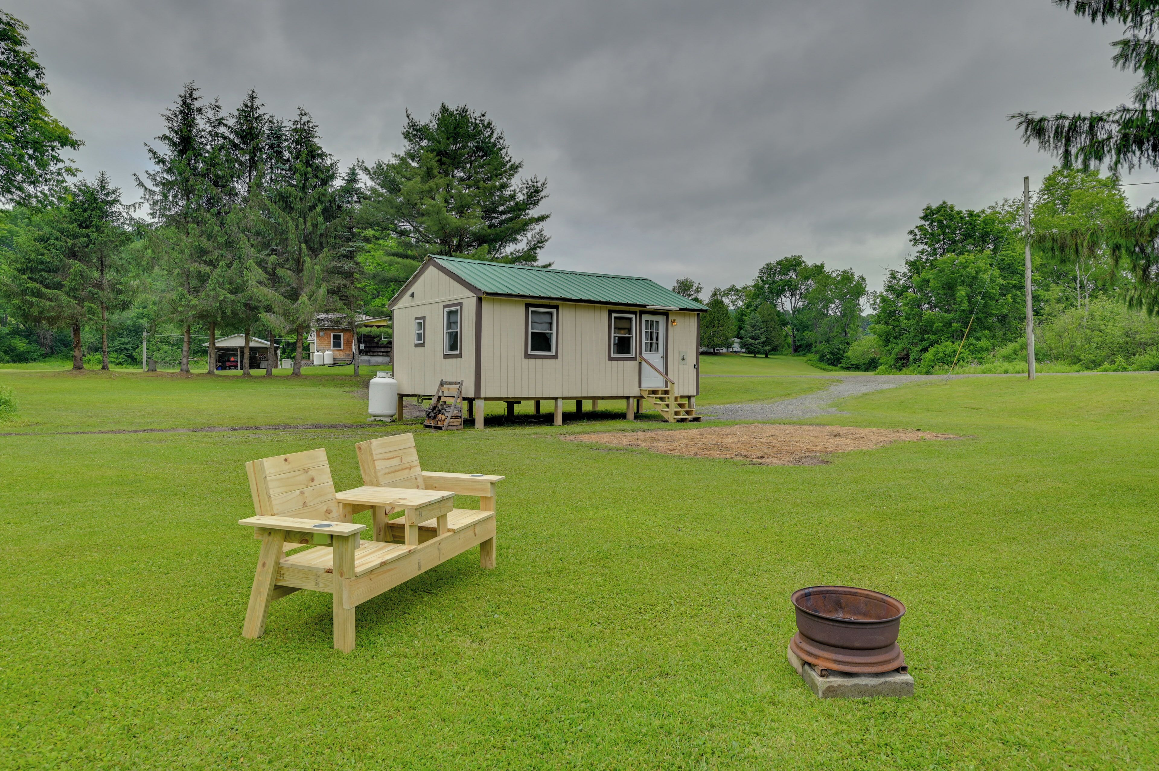 A small wooden house with a green roof, surrounded by a grassy field and trees. There are two wooden chairs and a small table in the foreground.