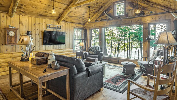A rustic cabin interior with wooden walls, a large window, a sofa, a television, and a wooden dining table. - Quiet Lakefront Conover Cabin Near ATV Trails (Conover)