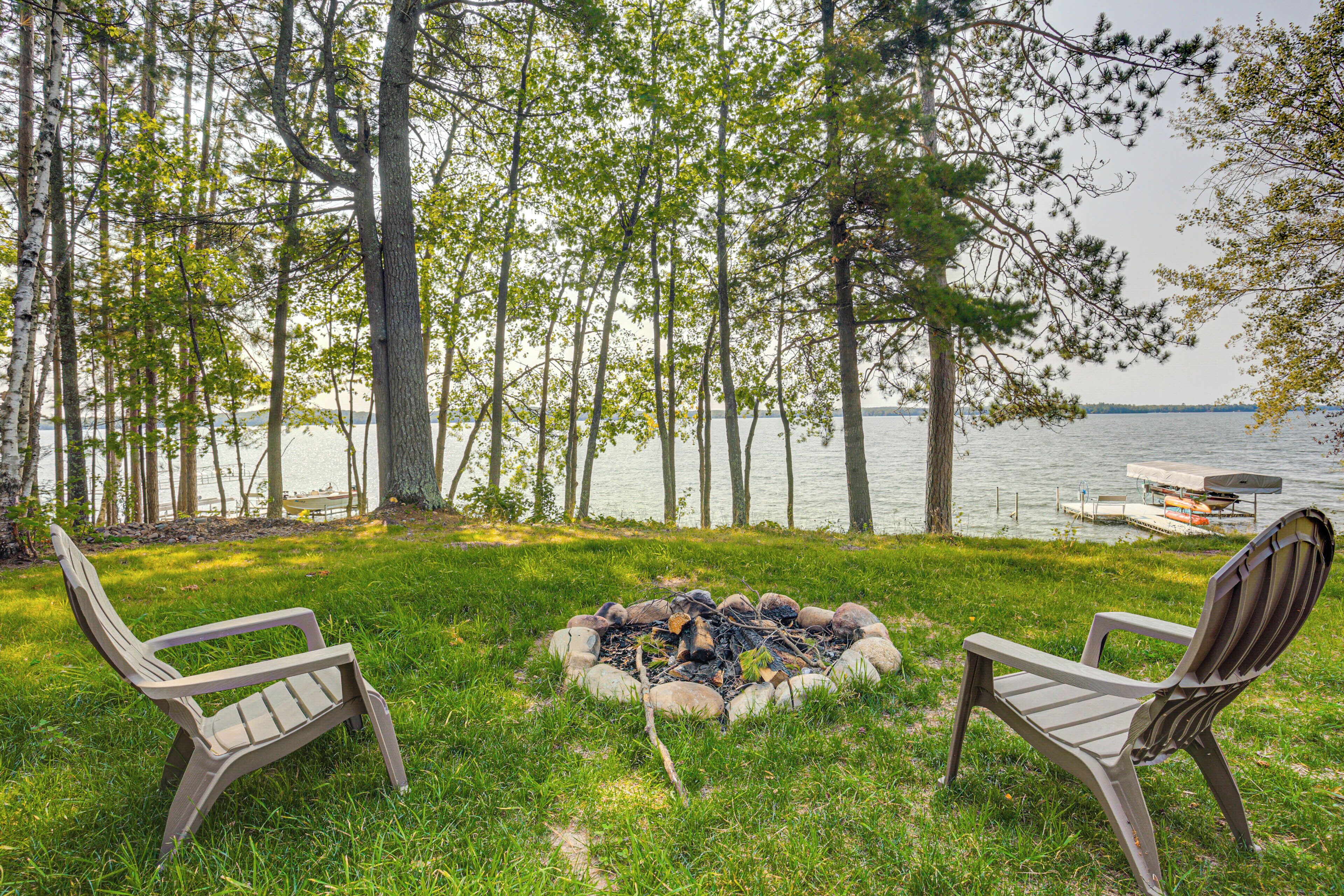 A lakeside view with two Adirondack chairs facing a fire pit surrounded by a grassy area and trees.