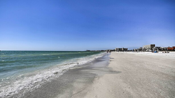 Ein weiter Sandstrand mit sanften Wellen, blauem Himmel und Gebäuden im Hintergrund.