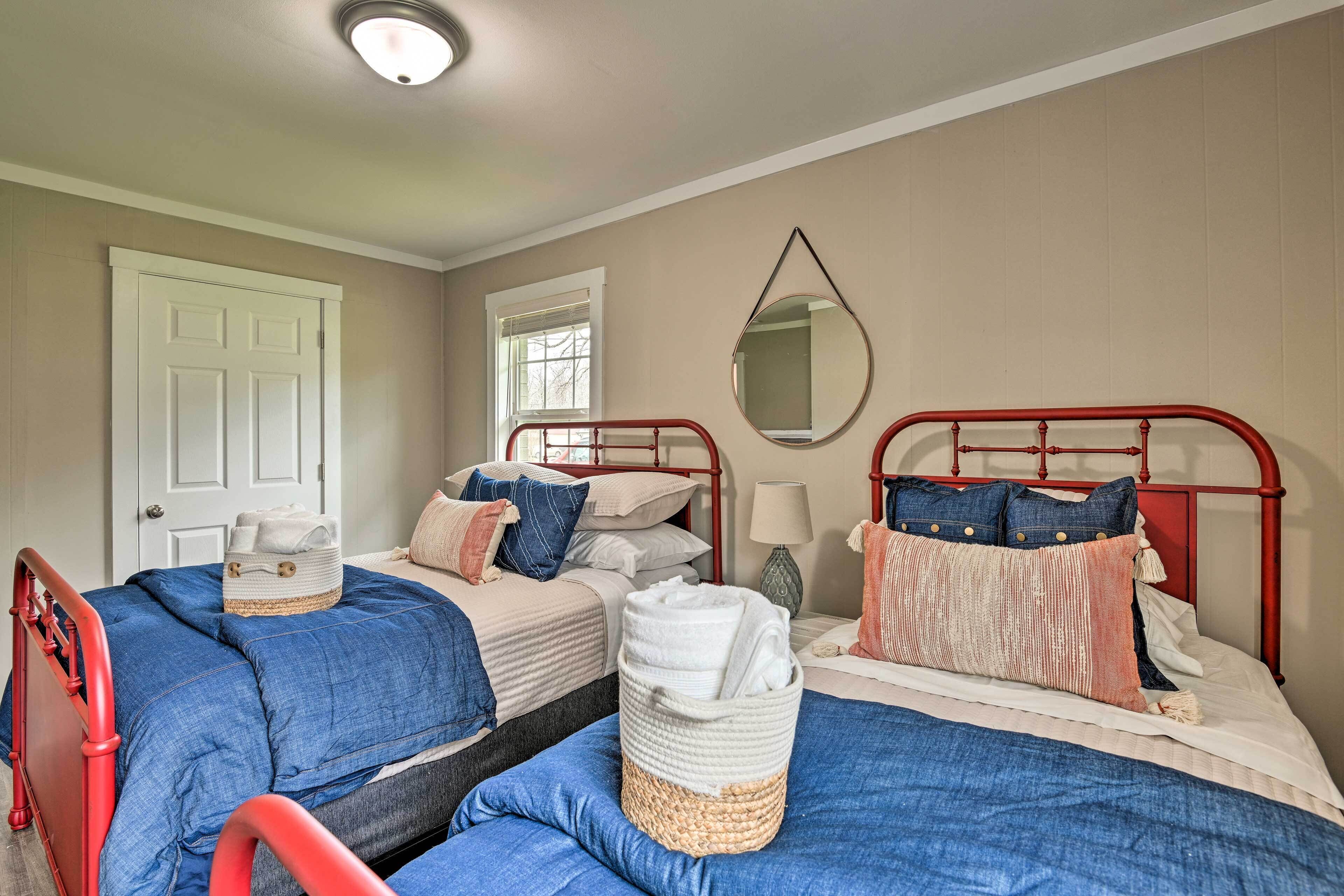 Quarto com duas camas, cada uma com estrutura metálica vermelha, roupa de cama azul e diversos travesseiros. Há uma porta branca, uma janela com persianas e um espelho na parede.